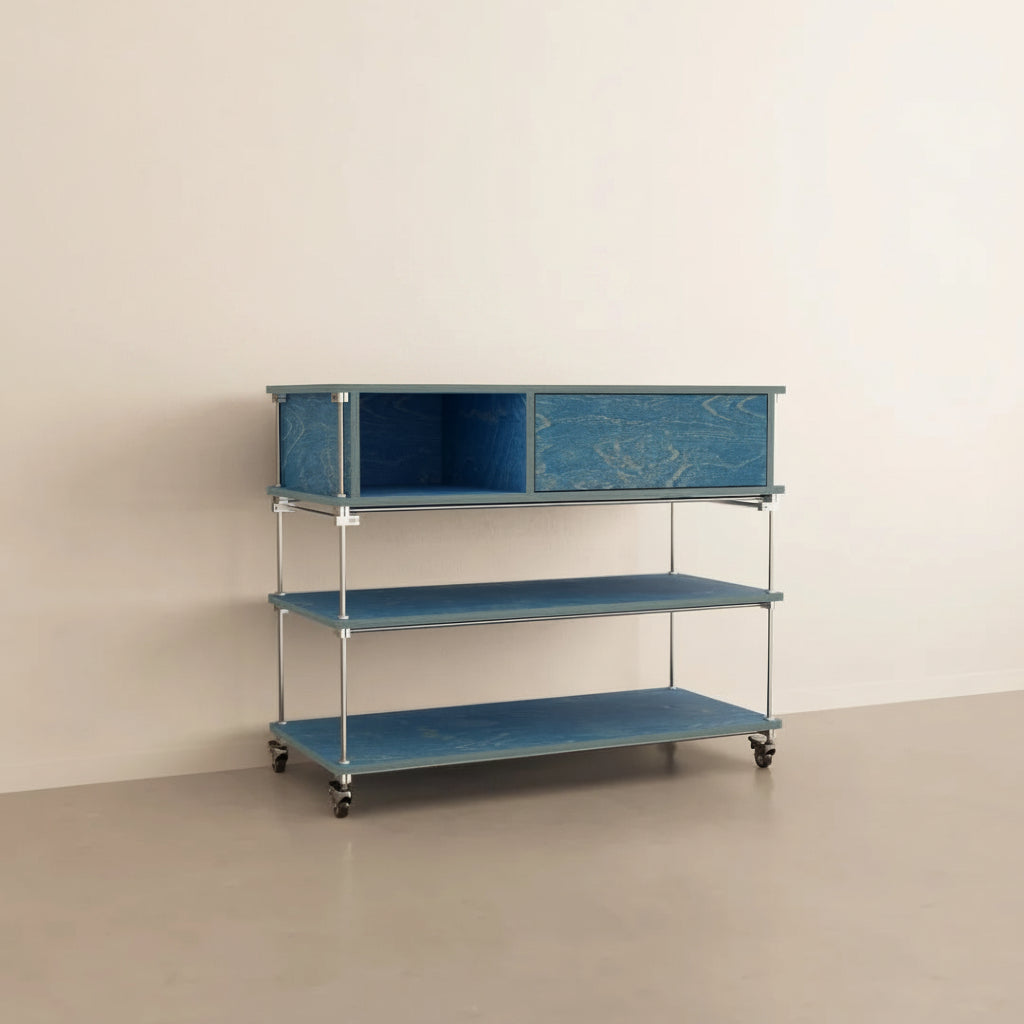 A blue three-tier storage cart with a metal frame and caster wheels, styled with a red desk lamp, a book, and red roses against a minimalist beige wall and floor.